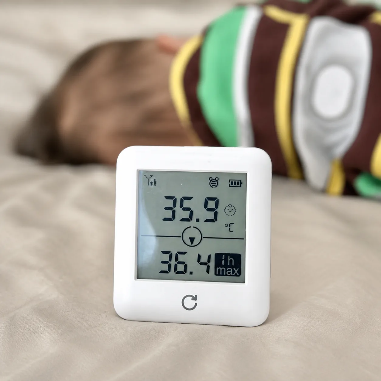 OnCorva bedside temperature monitor standing on a bed beside a sleeping baby, its screen showing 35.9 °C